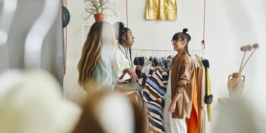 Three people are standing in a clothing store, engaged in conversation. They are surrounded by various garments on hangers and racks, including a striped dress one person is holding. The store has a minimalist decor with plants and hanging clothes.