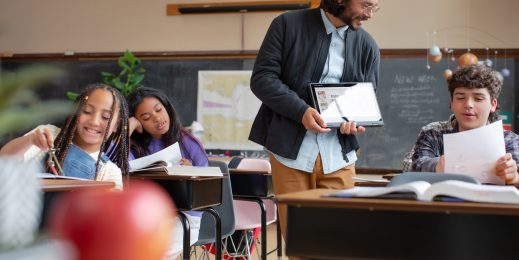 A classroom with students at desks, engaged in reading and writing. A teacher stands in the middle holding a tablet. The background includes a chalkboard with drawings and notes, and an apple on a desk in the foreground.