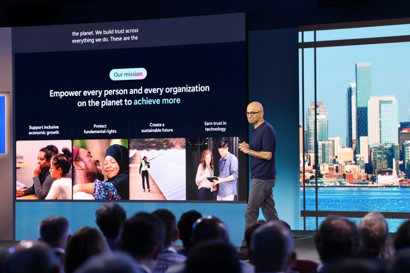 Man stands on stage with technology news projected in the background.