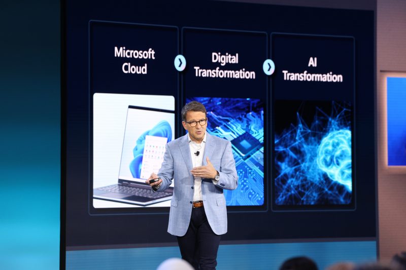 Man stands on stage with technology news projected in the background.