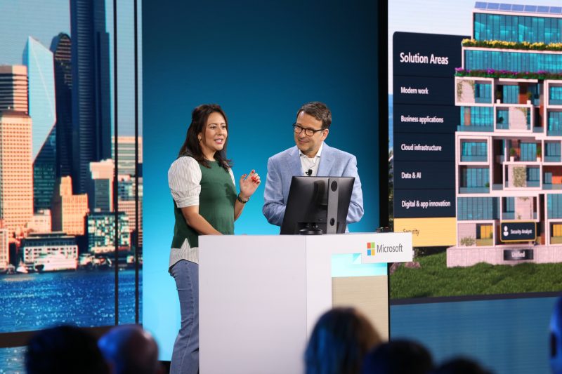 A man and a woman stand on stage behind a podium looking at a computer