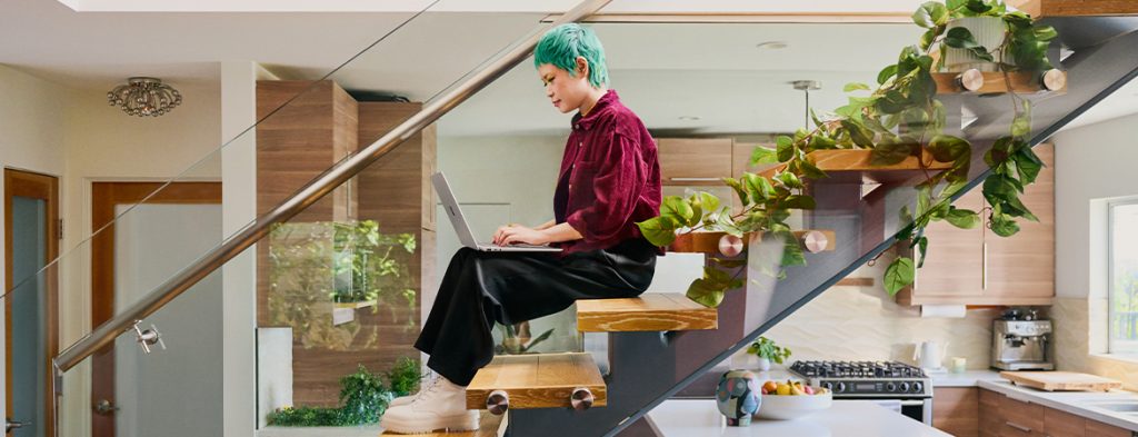 A person sits on stairs with potted plants on the steps by the kitchen, working on a laptop.