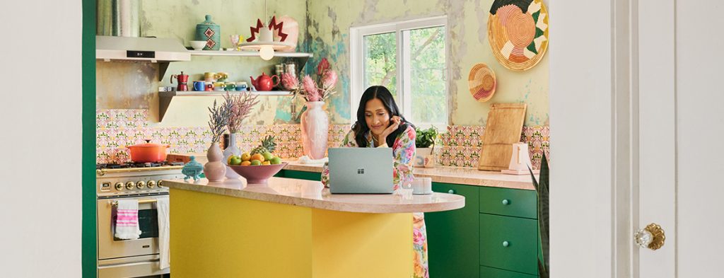 A woman stands at a kitchen island working on a laptop.