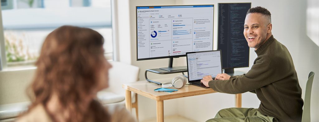 Two people work in an office, with one person working on a laptop with two external monitors.