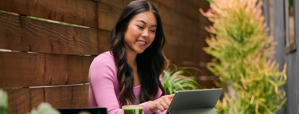 A woman sits outside by a wooden fence working on a Surface tablet.