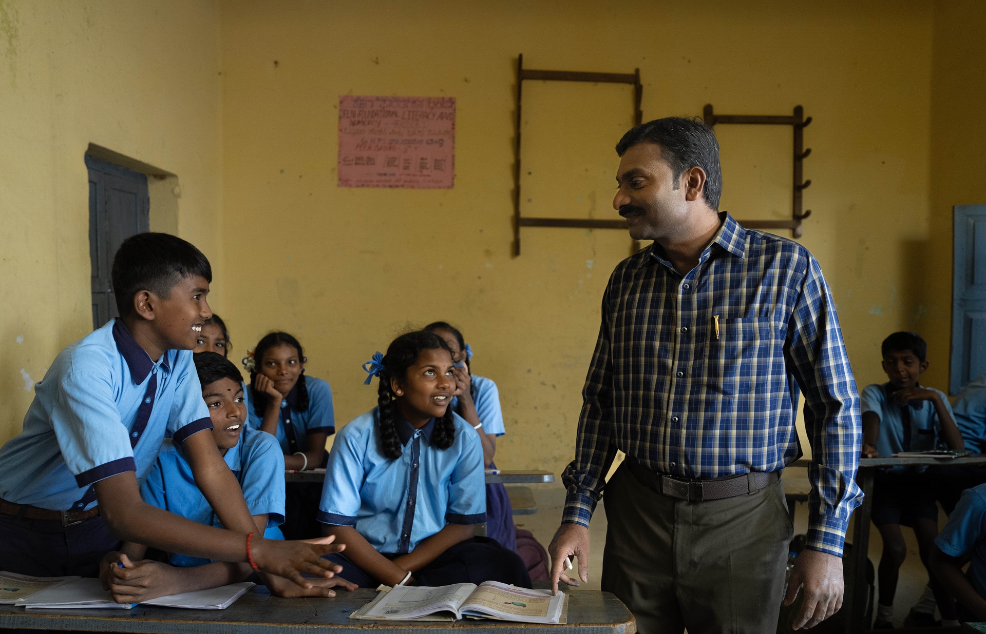 A male teacher in a blue plaid shirt interacting with students in blue uniforms in a classroom 