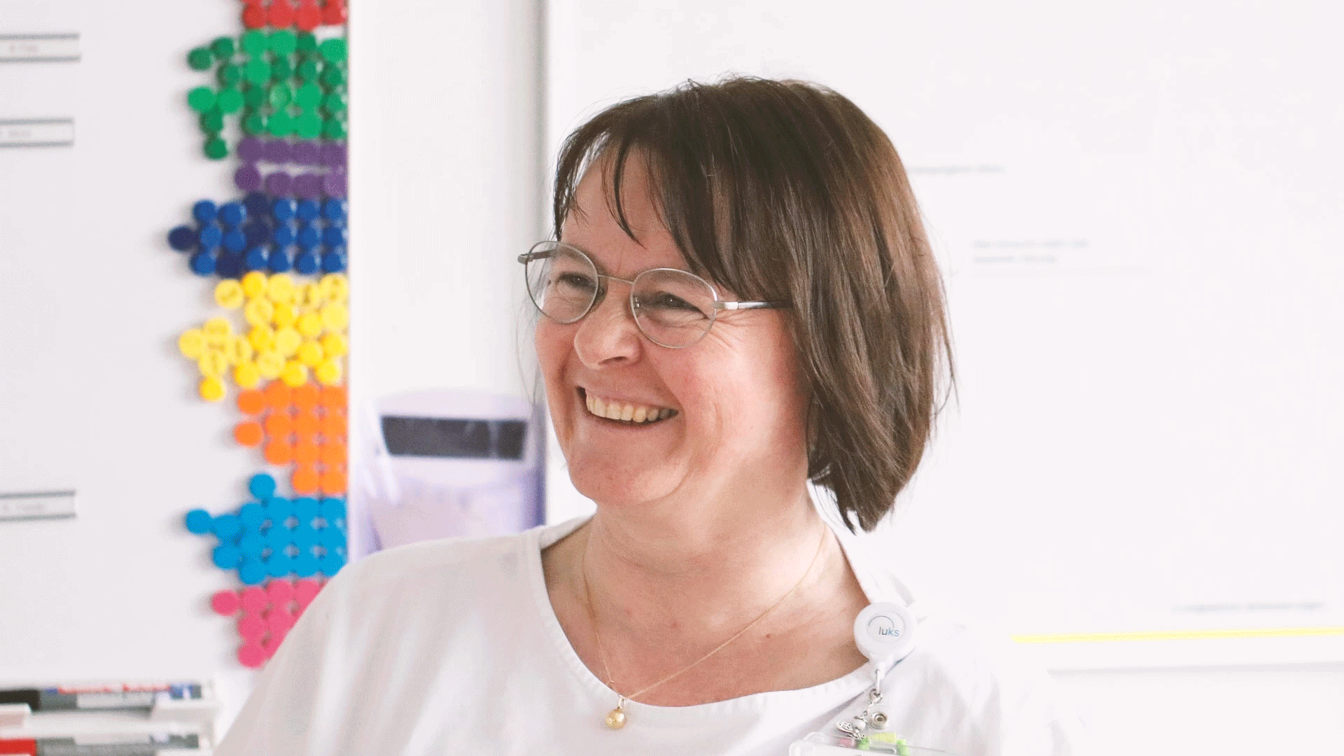 A smiling healthcare professional wearing glasses and a badge, standing in a brightly lit room with a colorful display in the background.