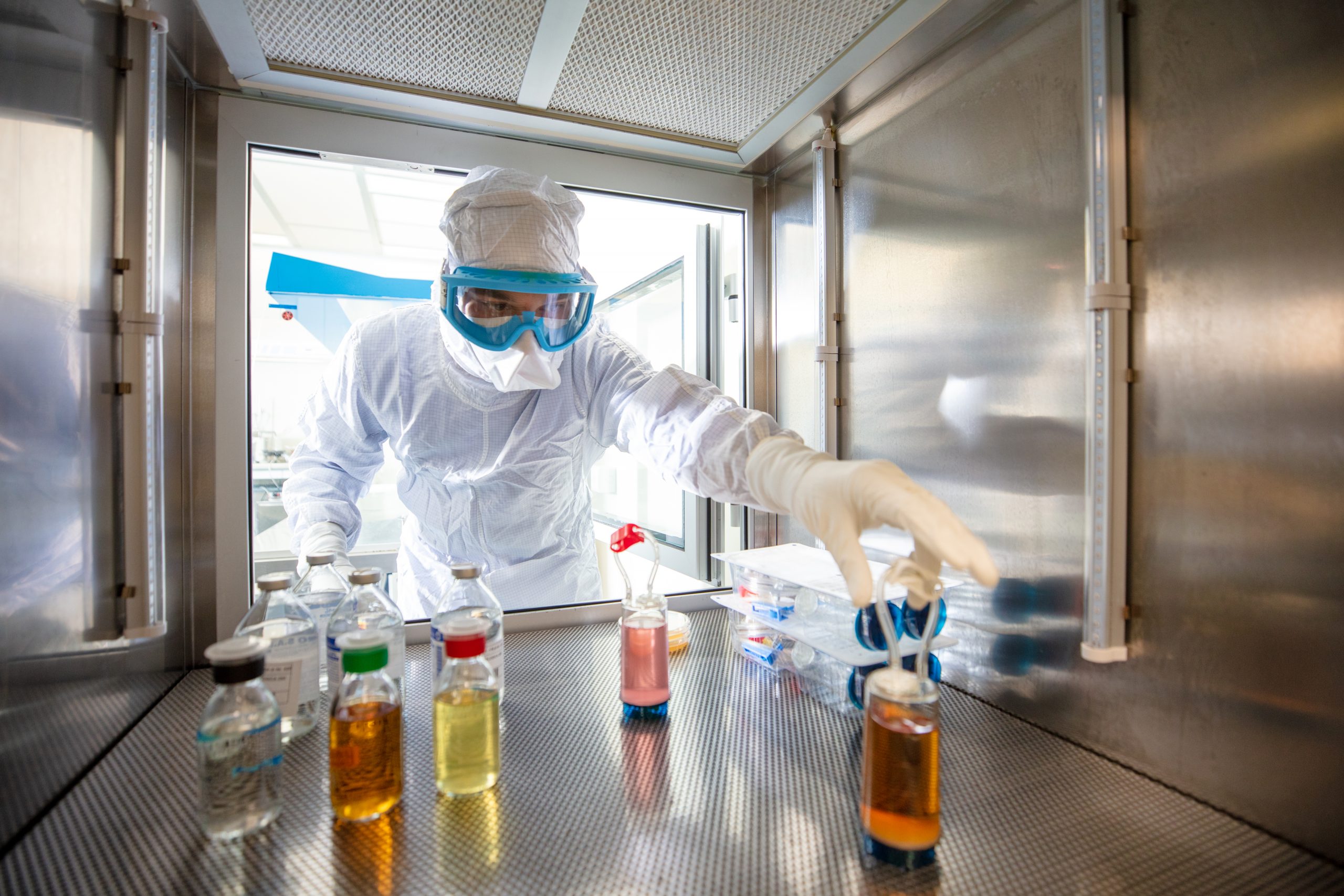 A person in full personal protective equipment (PPE), including a face mask, goggles, gloves, and a lab suit, working in a controlled laboratory environment. The individual is handling various liquid-filled vials.