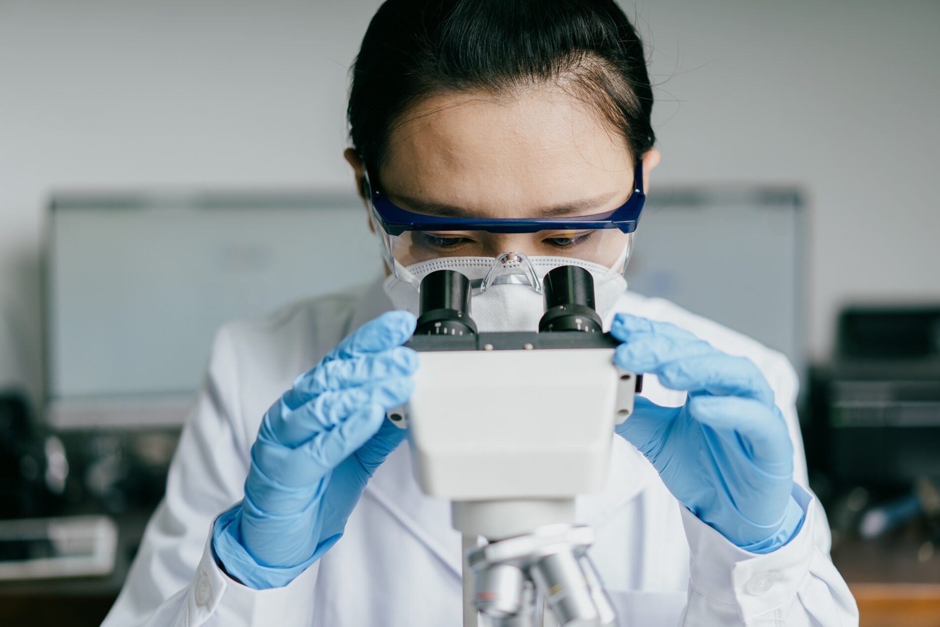 A person wears safety goggles, a face mask, and blue gloves, closely examining a sample through a microscope in a laboratory.