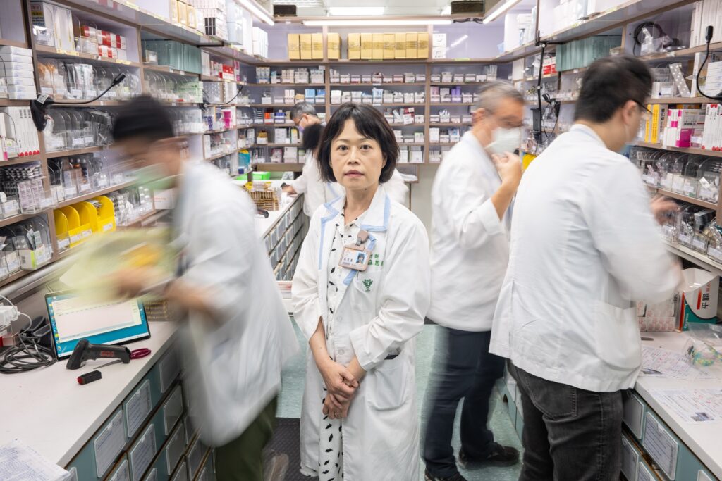 A healthcare professional standing in the middle of a busy hospital pharmacy, surrounded by other staff members working quickly in the background.