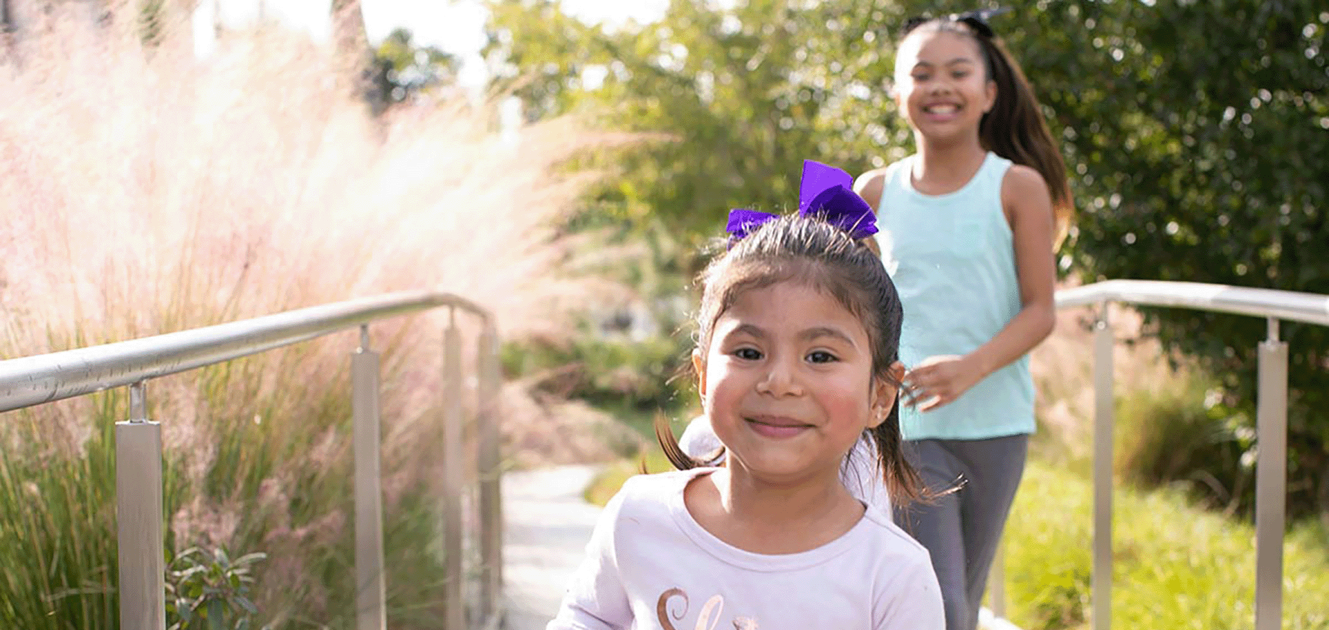 Two young children smiling and running outdoors on a pathway, surrounded by greenery and sunlight. The child in front has a purple bow in her hair, and the background is blurred slightly, giving a sense of motion and joy.