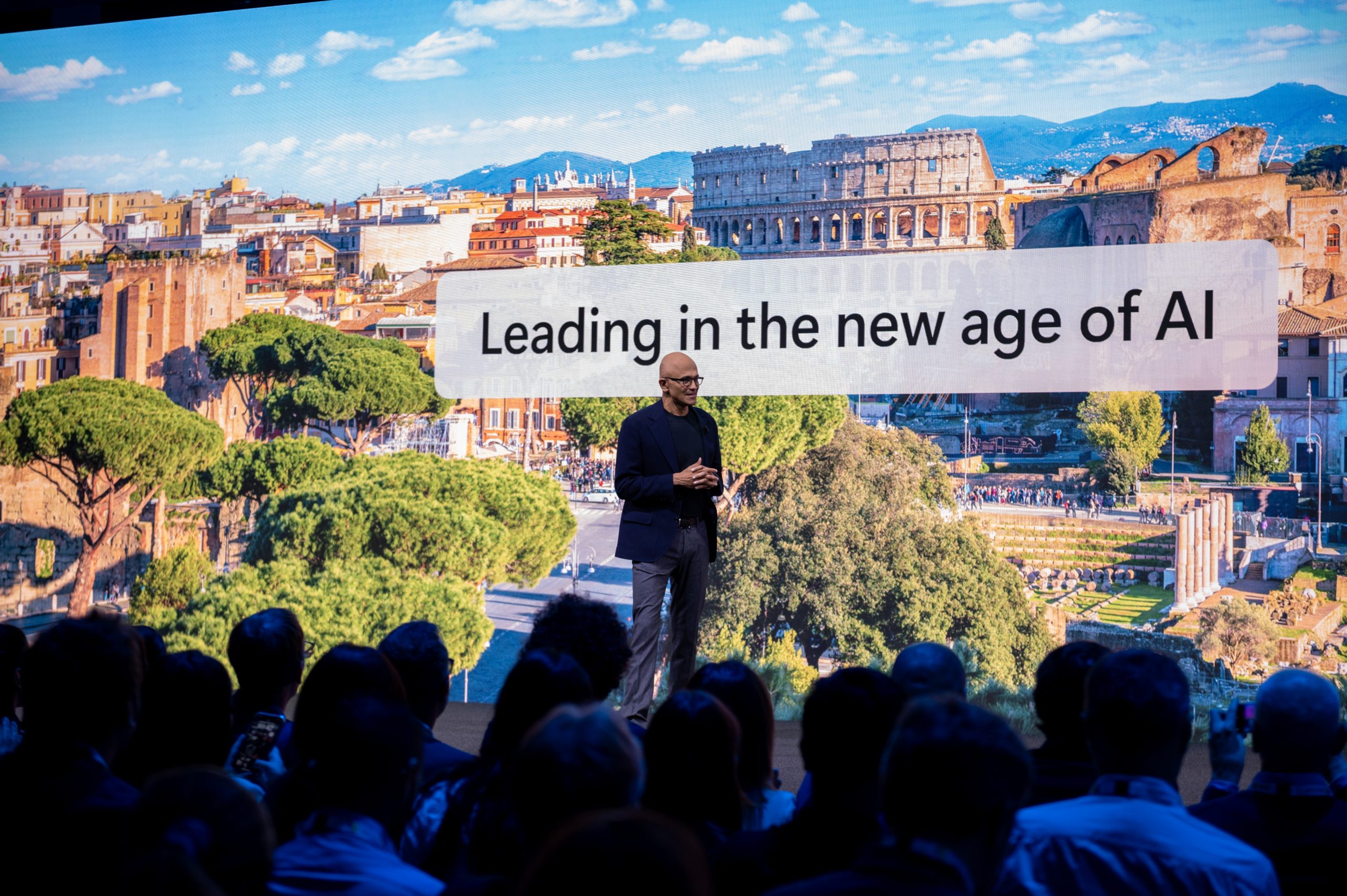 Satya Nadella is standing onstage with a presentation behind him. The image is a landscape of Rome and the words read 'Leading in the new age of AI'. There are silhouettes of audience members as they watch Satya present.