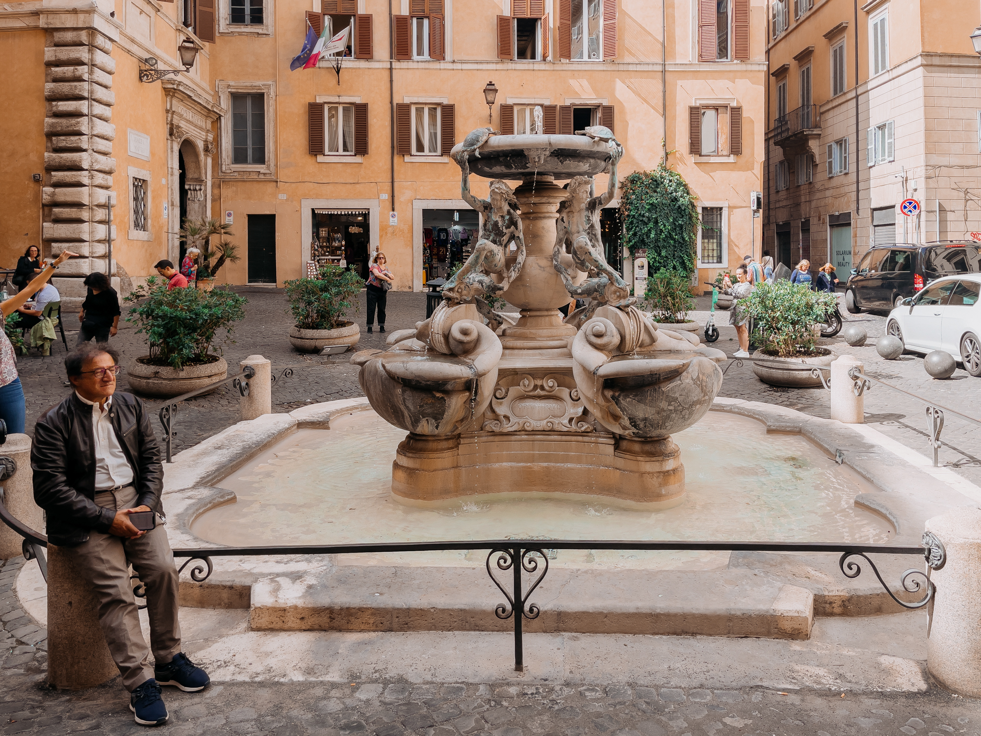 A man sitting on a low iron barrier surrounding an ornate fountain. He is wearing a white shirt and brown leather jacket.