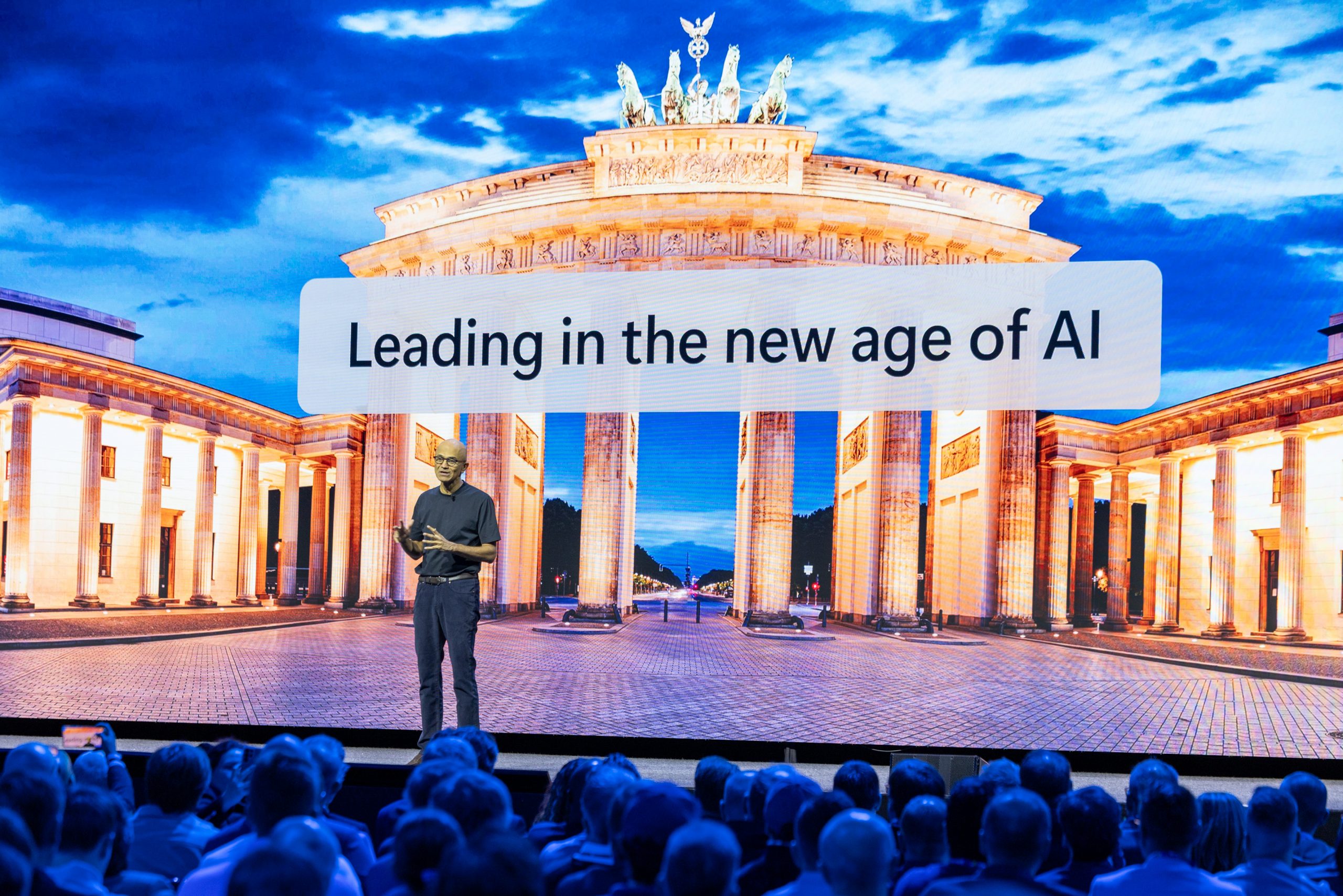 Satya Nadella stands onstage giving a presentation. He is wearing a black top and dark trousers. Behind him is a large screen displaying an image of the Brandenburg Gate. Words on the screen say "Leading in the nw age of AI"