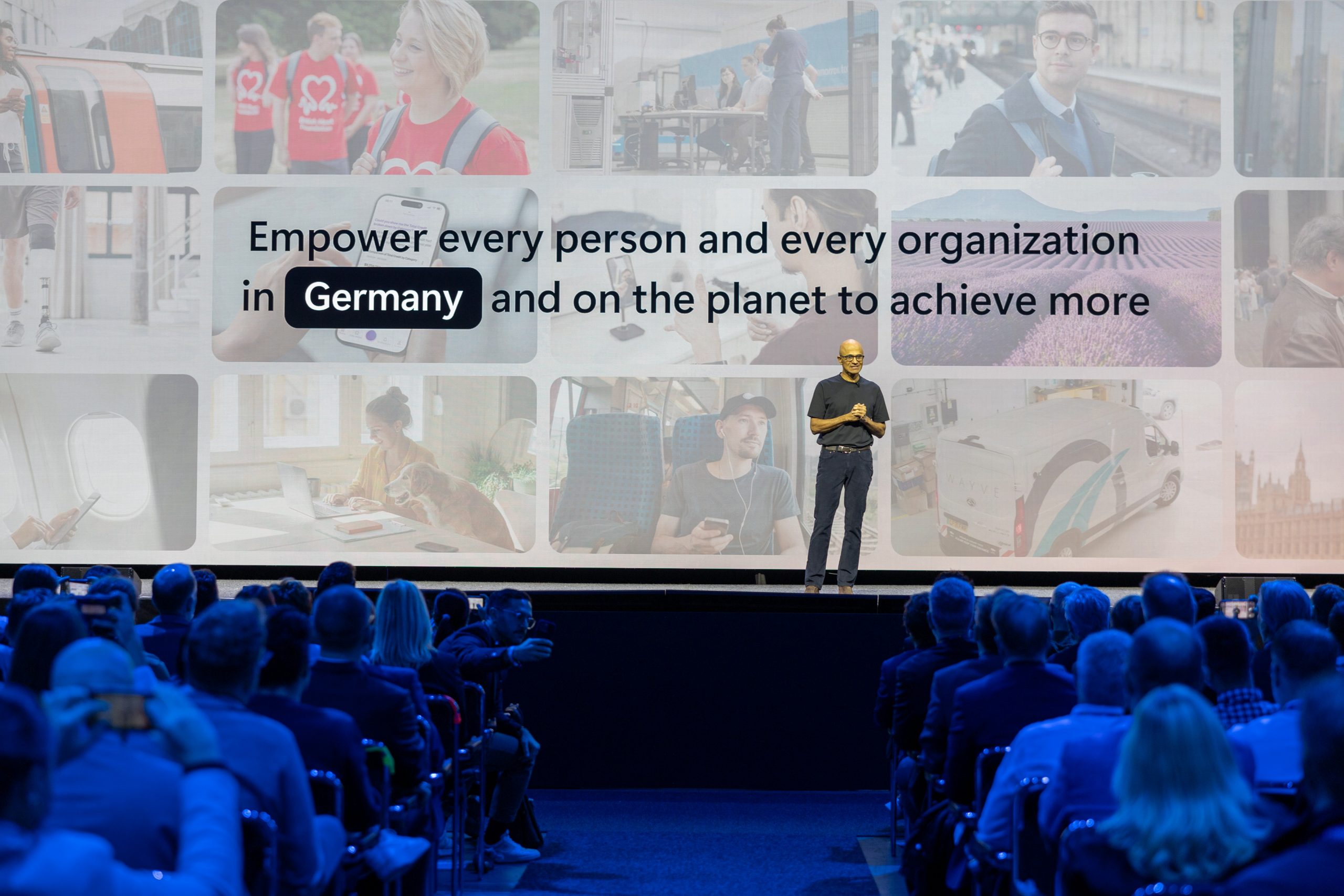 Satya Nadella stands onstage giving a presentation. He is wearing a black top and dark trousers. Behind him is a screen of different photos depicting how technology is helping people. The company mission statement has been adapted to say: ‘Empower every person and every organization in Germany and on the planet to achieve more.