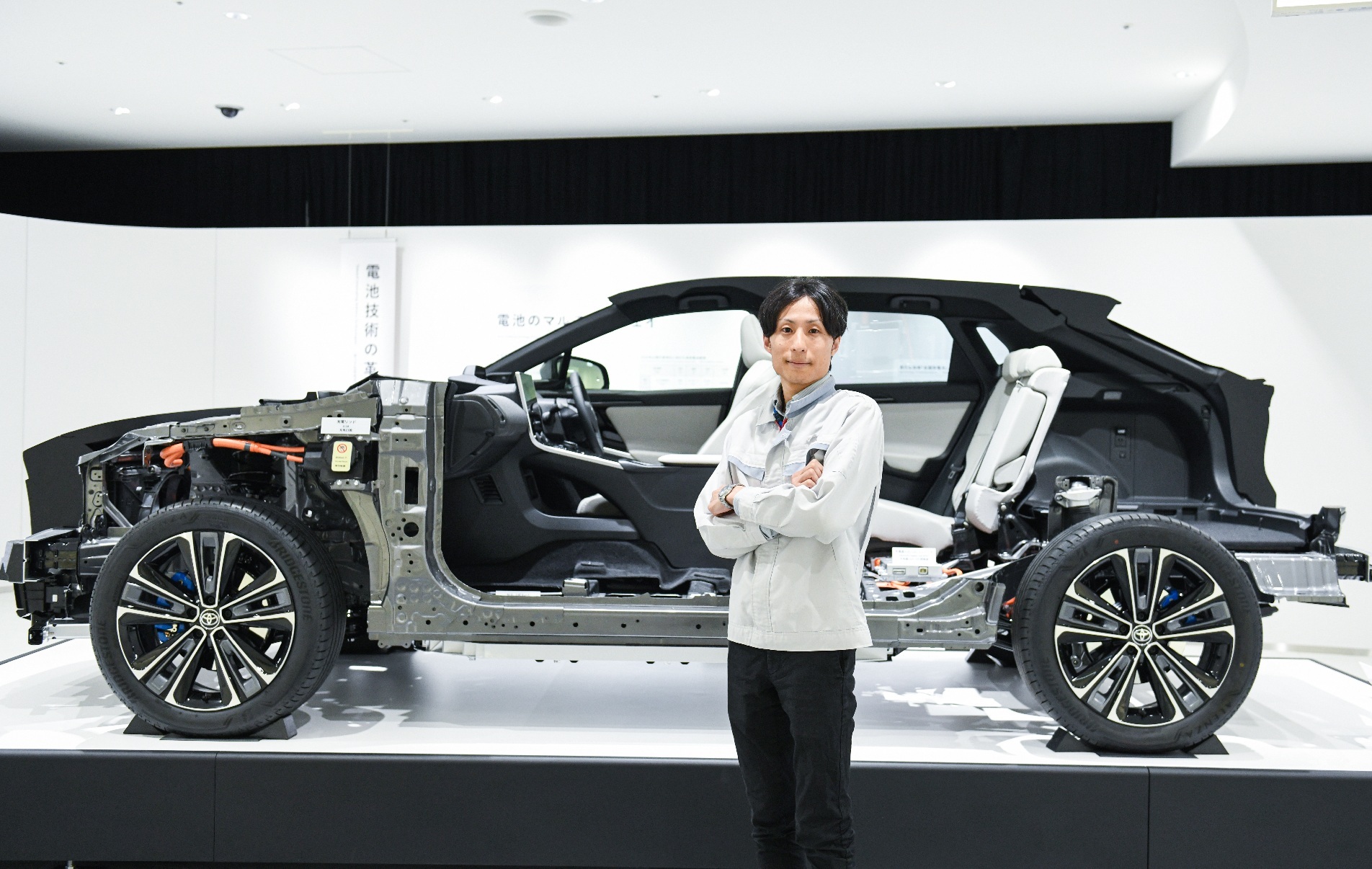 A man standing with his arms crossed next to a car display with the vehicle body peeled off, showing the engine and other internal parts. 