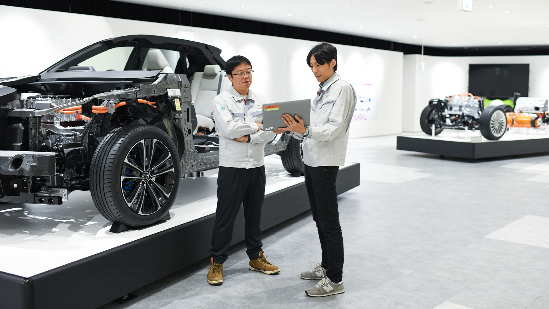 Two men look at a laptop next to a car display with the vehicle body peeled off to show the engine and other internal components.