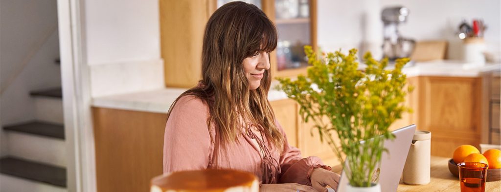 Uma mulher senta-se a uma mesa perto de uma cozinha a trabalhar num computador portátil