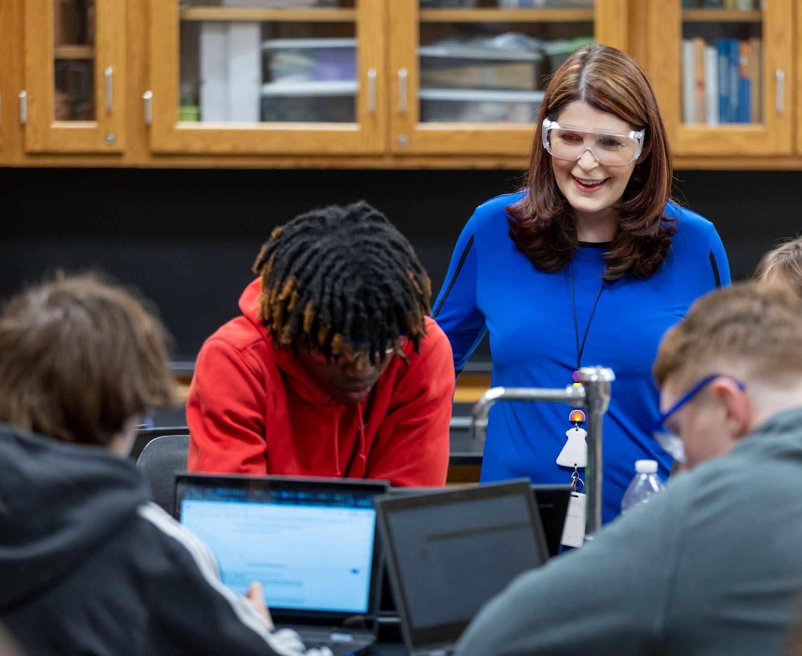 A student in a red hoodie is focused on the laptop screen, while a woman, teacher in a blue shirt stands nearby, observing.