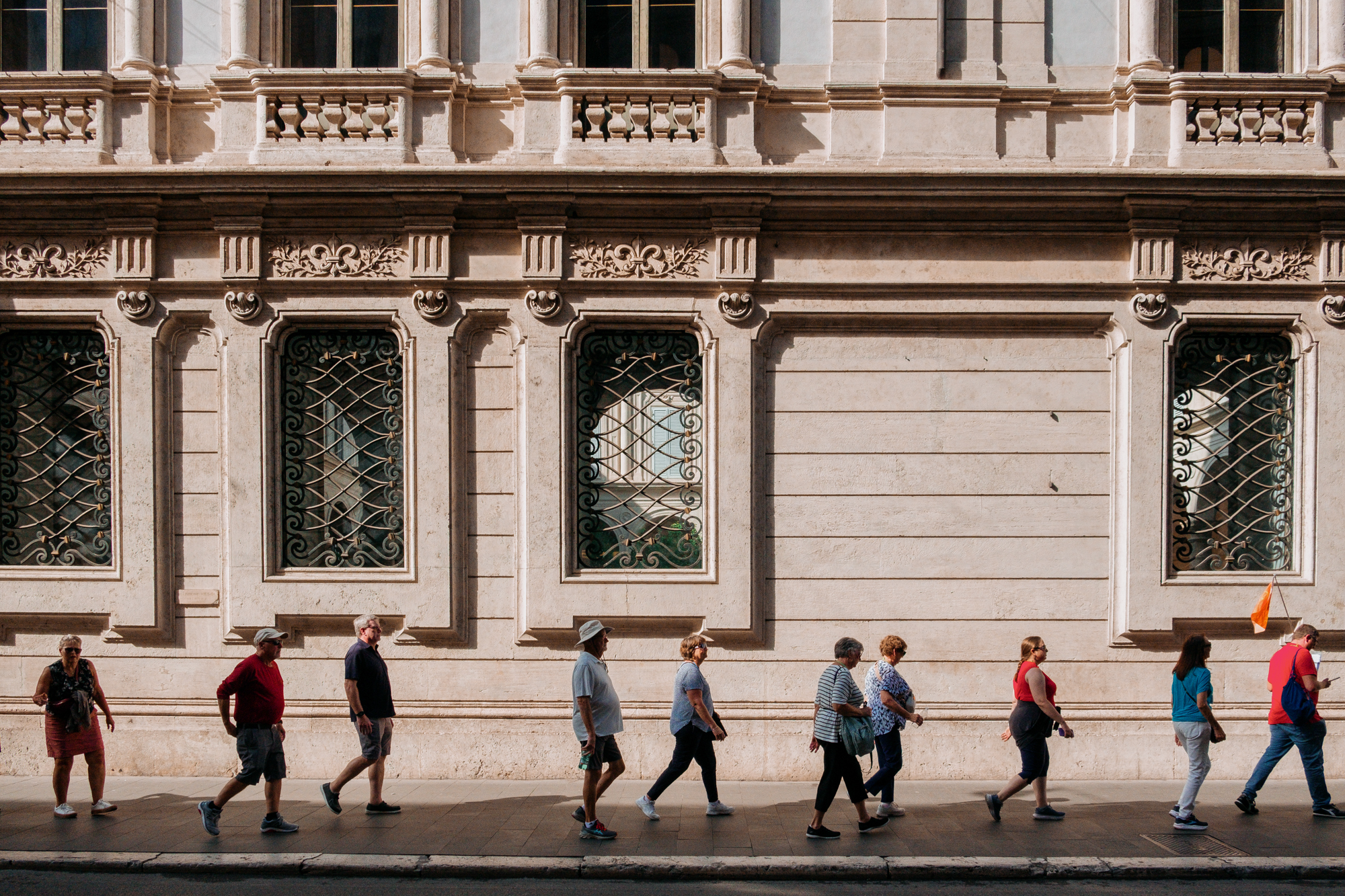 A line of tourists, following a leader with a flag, on a city sidewalk with an ornate building behind them